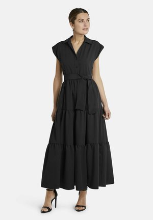 Frau trägt ein ärmelloses, schwarzes Maxikleid mit Stufen, Kragen, Taillengürtel und schwarzen Sandalen mit hohem Absatz, steht und schaut seitlich.