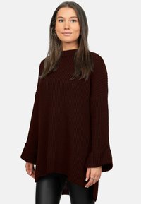 Maglione oversize bordeaux in maglia con texture a coste, maniche larghe e orlo asimmetrico. Abbinato a pantaloni neri aderenti per creare un contrasto.