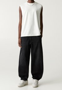 Top blanco sin mangas con cuello redondo, combinado con pantalones vaqueros negros de corte amplio y zapatillas blancas, con detalles de costuras visibles.