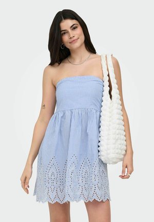 Ung kvinde iført en blå, skulderløs kjole med striber og hulmønster, der holder en hvid skuldertaske med struktur mod en ensfarvet baggrund.