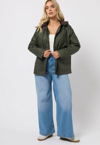 Veste vert olive avec un revêtement de col marron, fermeture par boutons et zip, deux poches avant, portée avec un jean bleu clair à pattes larges et des chaussures beiges.