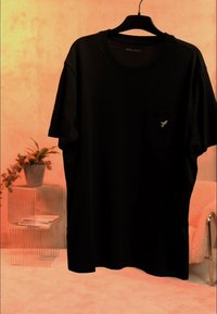 T-shirt noir à manches courtes avec petit emblème d'oiseau sur la poche, suspendu à un cintre noir dans une pièce aux tons chauds avec une plante et une chaise en arrière-plan.