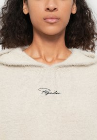 Gebreide hoodie in lichtbeige met een zachte textuur. Heeft een capuchon met trekkoord en een zwart geborduurd logo op de borst.