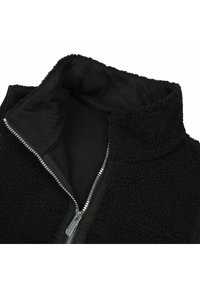 Veste en polaire noire avec col montant et fermeture éclair argentée partiellement ouverte laissant apparaître une doublure intérieure douce.