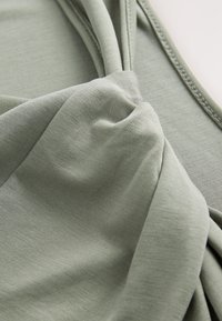 Haut en tissu vert avec un détail de nœud torsadé au centre. Le matériau a une texture lisse et un léger effet stretch, présentant une teinte douce et tamisée.