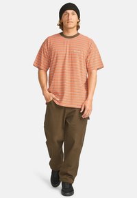 Stribet t-shirt i orange og hvid med en mørkebrun rund halsudskæring. Matcher med løstsiddende olivengrønne bukser og sorte sneakers.