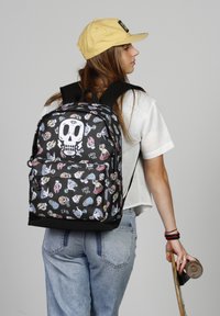 Mochila negra con patrones coloridos de calaveras y temática de patinaje; cuenta con correas acolchadas y un bolsillo frontal. La tela parece duradera y texturizada.