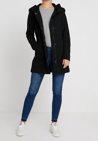 Black hooded coat with a textured pattern, front zipper, and snap buttons. Worn over a grey top, paired with blue jeans and white sneakers.