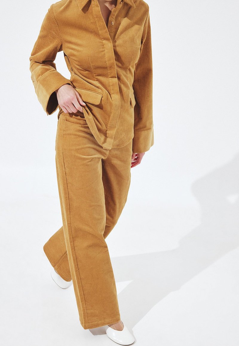 Person wearing mustard yellow corduroy suit with flared pants and jacket, adjusting jacket hem, standing on white floor with white shoes.