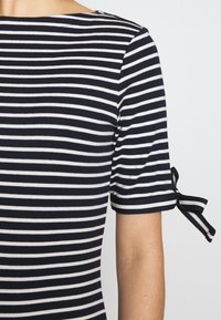 Camisa de manga corta a rayas horizontales en blanco y negro con un detalle de lazo atado en la manga, llevada en un brazo descubierto contra un fondo plano.