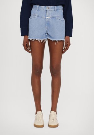 Personne portant un short en jean bleu clair avec des ourlets effilochés, un haut à manches longues foncé et des baskets blanches, debout devant un fond uni.