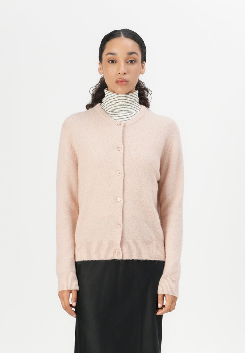 Light pink cardigan made of soft knit material. Features a round neckline, five buttons, ribbed cuffs, and hem. Worn over a striped turtleneck.