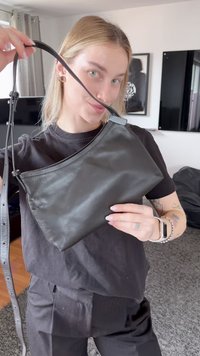 Persona joven con cabello rubio sosteniendo la correa de un bolso de cuero negro cerca de su cara, de pie en una sala de estar.