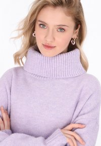 Pullover turtleneck de culoare mov deschis, cu guler și manșete cu nervuri, realizat dintr-un material moale; modelul își încrucișează brațele pe un fundal neutru.
