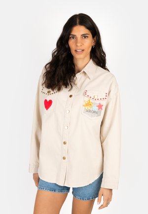 Woman with long dark hair wears beige buttoned shirt decorated with red heart, yellow flower, star, and "AMORE" text, paired with denim shorts.