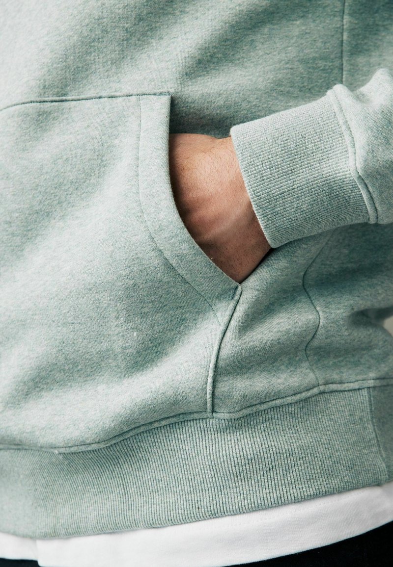Mintgroene hoodie met een kangeroezak, geribbelde manchetten en zoom. Materiaal lijkt zacht te zijn, met een gestructureerde afwerking. Draaglaag over een wit shirt.