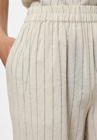 Pantaloni in lino color beige chiaro con sottili righe verticali nere, dotati di una cintura elastica e tasche laterali per maggiore comodità.