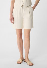 Beige tailored shorts with pleats, made from textured fabric, featuring side pockets. Paired with tan slide sandals with woven straps.