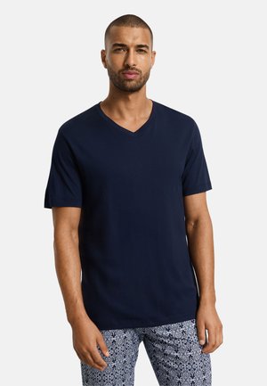 Mies, jolla on lyhyet hiukset ja parta, pukeutunut tummansinisään V-kaulaiseen t-paitaan ja kuvioituihin sinivalkoisiin housuihin, seisomassa yksivärisen taustan edessä.