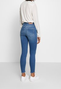 Jeans de mezclilla azul de tiro alto con un ajuste ceñido, un suave desvanecimiento, dobladillo deshilachado y bolsillos traseros. Combinados con una blusa crema y zapatillas blancas.