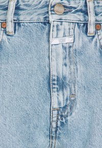 Jeans de denim azul claro con cintura alta, cierre de botón de cobre y un sutil detalle desgastado con acentos de costura naranja.