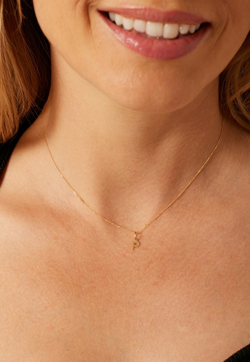 Collier ton or avec une chaîne fine et un petit pendentif en forme de lettre « P », reposant sur la peau au niveau du cou. Design simple et délicat.