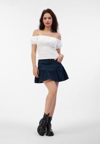 Weiße schulterfreie Bluse aus strukturiertem Stoff, kombiniert mit einem dunklen Denim-Röckchen mit Schlag. Das Model trägt schwarze Knöchelstiefel mit einer dicken Sohle.