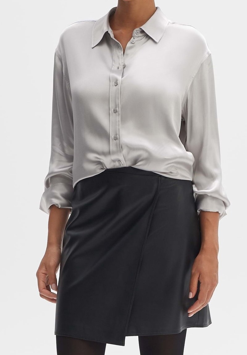Femme portant un chemisier en satin gris clair boutonné avec manches retroussées et une jupe portefeuille noire sur des collants noirs, debout devant un fond blanc.