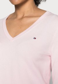 Jasnoróżowy sweter z dekoltem w serek, wykonany z miękkiego materiału, z małą naszywką z logo w czerwonych, białych i niebieskich akcentach na piersi.