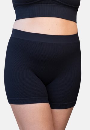 Frau trägt schwarze, gerippte Biker-Shorts mit hohem Bund und passendes Crop-Top, zeigt Bauch und Oberschenkel vor einem einfarbigen Hintergrund.