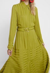 Robe à manches longues en tissu vert vibrant avec une texture rayée, dotée d'un col, d'une taille ceinturée avec une boucle circulaire et d'une jupe évasée.