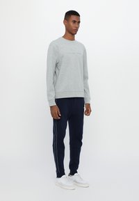 Sudadera gris con logo bordado, combinada con joggers marinos que cuentan con franjas laterales blancas. El modelo lleva zapatillas blancas. Look simple y casual.