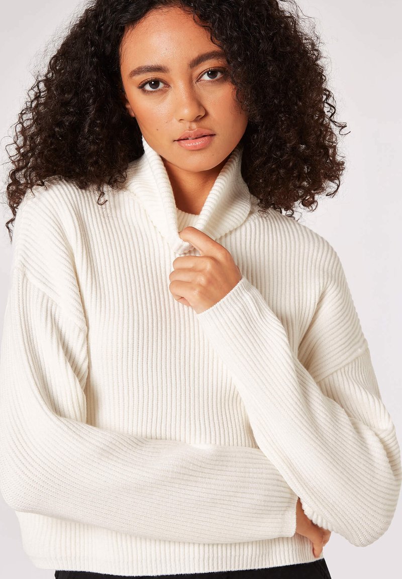 Maglione a coste color crema con colletto alto e piegato e maniche lunghe e morbide, caratterizzato da un fit rilassato e una texture morbida.