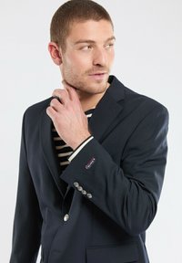 Blazer bleu marine à coupe ajustée, design à simple boutonnage, boutons argentés, et chemise à rayures contrastées en dessous. Texture de tissu lisse.