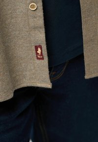 Brūns zigzagains krekls ar pogātu priekšu un teksturētu apdari; ar bordo logotipa uzlikumu apakšmalā, kombinēts ar tumšām džinsām.