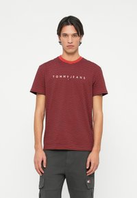 Rød og marineblå stripete T-skjorte med rund hals og kontrasterende rød krage. Har hvit "TOMMY JEANS" logo over brystet.