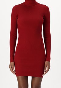 Abito a maniche lunghe con collo alto, di colore rosso e a coste, dal design aderente e l'orlo che arriva a metà coscia. Include un piccolo etichetta con logo sull'orlo.