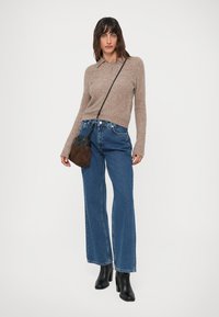 Mujer vestida con un suéter de punto beige, jeans de mezclilla azules, botas de tacón negras y un bolso bandolera marrón-negro sobre un fondo blanco.