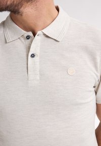 Kortärmad polotröja i ljus beige tyg, med en klassisk krage, två marinblå knappar och en liten rund logotyp på vänster bröst.