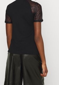 Černé tričko s krátkými krajkovými rukávy a zaobleným lemem, spárované s lesklými tmavě zelenými šortkami s hladkou texturou.