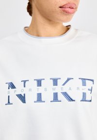 Bijela dukserica s plavim vezom "NIKE" i "SPORTSWEAR". Ima okrugli izrez i meku, teksturiranu tkaninu.