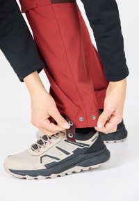 Bottines impermeables beiges avec semelle noire et accents gris, associées à un pantalon rouge avec des fermetures à pression au bas, des mains ajustant l'ajustement.