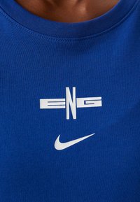 Lyhythihainen sininen T-paita, joka on valmistettu puuvillasta. Paidassa on valkoiset vaakaraitat ja "ENG"-logo. Alareunassa on valkoinen Nike-swoosh-logo.