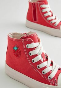 Czerwone płócienne buty typu high-top z białymi oczkami i sznurówkami. Posiadają haftowany wzór truskawki po boku. Teksturalny materiał i szyte akcenty.