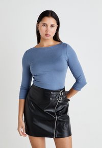 Μπλε μπλούζα με μακριά μανίκια σε απαλό ύφασμα, συνδυασμένη με μαύρη μίνι φούστα από συνθετικό δέρμα που διαθέτει μπροστινό φερμουάρ και δύο διακοσμητικά αγκράφες.