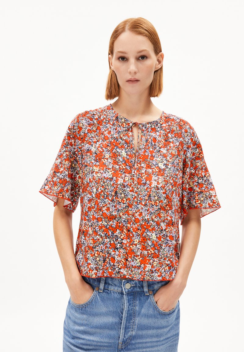 Kurzärmlige Bluse, leuchtend rotes Blumenmuster, leichtes Material, runder Halsausschnitt mit Bindedetail, lockere Passform. Kombiniert mit blauen Jeans.