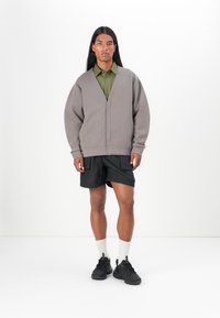 Grå oversized sweatshirt med V-ringning, bärs över en grön skjorta med knappar. Matchad med svarta shorts och svarta sneakers. Vita vadstrumpor.