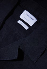 Chemise en coton noire avec col à boutons et étiquette de marque visible. Tissu texturé avec une finition soignée et des détails de design simples.