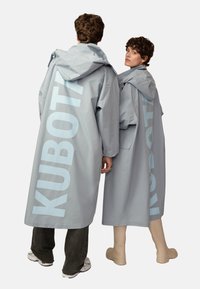Hellgraue, übergroße Regenmäntel mit Kapuze, versehen mit großen "KUBOTA"-Schriftzug in Hellblau auf dem Rücken und seitlichen Taschen. Glatte Textur.