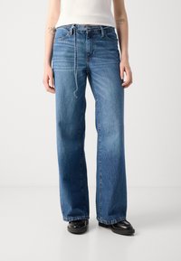 Jean bleu à jambe large en denim, avec taille haute, passants de ceinture, poches avant et ourlet effiloché. Des détails cousus sont visibles.